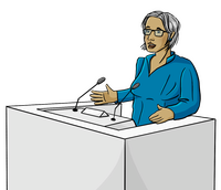 Eine Frau hat weißes Haar und eine blaue Bluse. Sie steht hinter einem weißen Podest. Auf dem Podest sind Mikrofone. Die Person erzählt etwas. Die Person hat eine dunkle Haut-Farbe. Die Person trägt eine Brille.